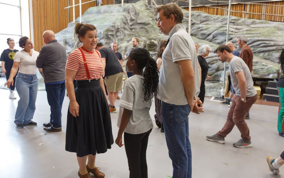 Katie Bird in a red and white striped top, red braces and black skirt speaks to a young girl standing in front of Ed Bennett wearing a casual grey top and blue jeans.