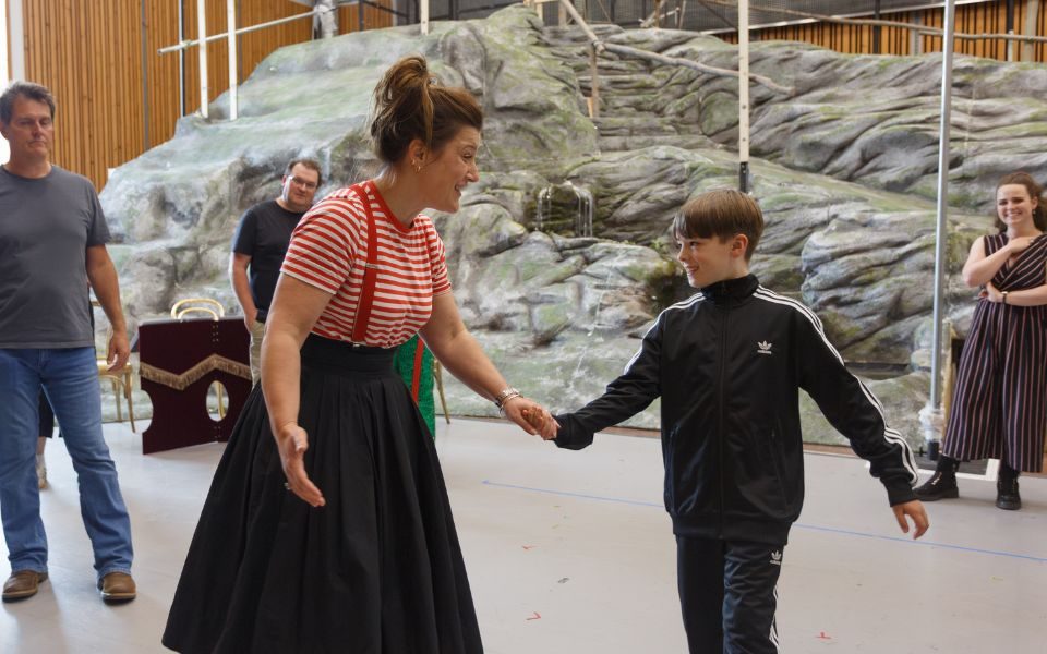 Katie Bird dressed in a red and white striped top, red braces and black skirt holding hands with Lucas dressed in a black sports jacket.