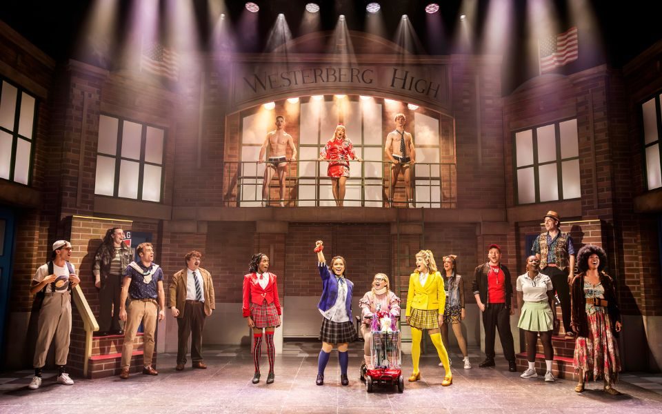 On stage in front of a high school set, a group of actors sing together in colourful costumes, including a character in a neck brace on a mobility scooter.
