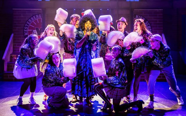 An ensemble of dancers hold fanned out open books around a central figure singing on stage.