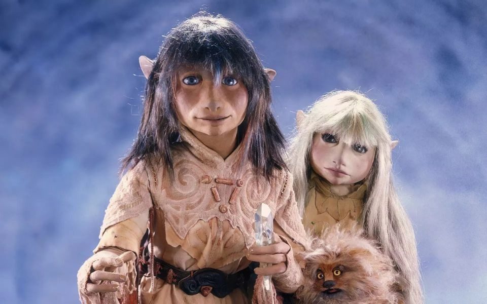 Two puppets of elf-like characters with long hair, in earthy, fantastical clothing and holding a translucent crystal.