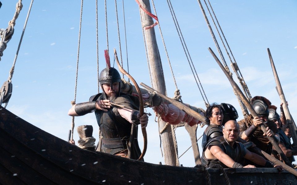 On a long wooden boat, a soldier in ancient armour points a bow and arrow, along with a group of other men holding large wooden oars.
