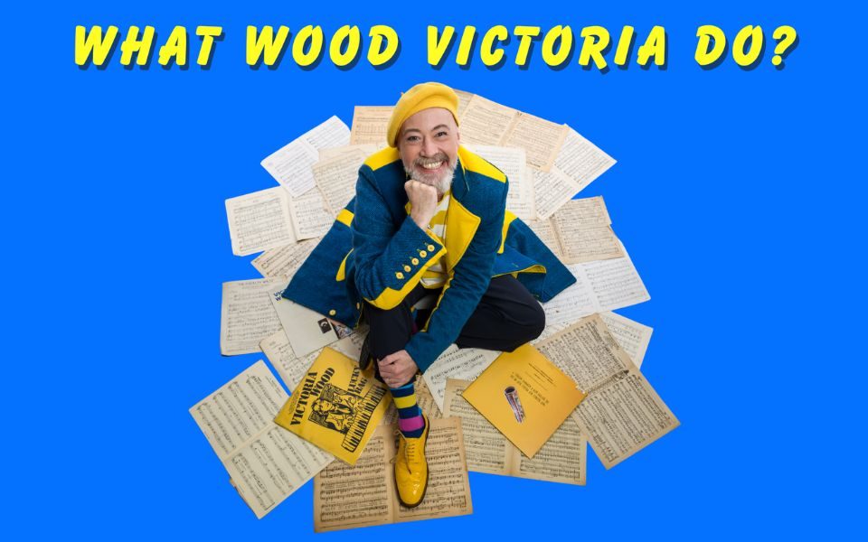 A man with a yellow beret and matching shoes sits smiling on pages of sheet music and Victoria Wood records. Text reads: What Wood Victoria Do?