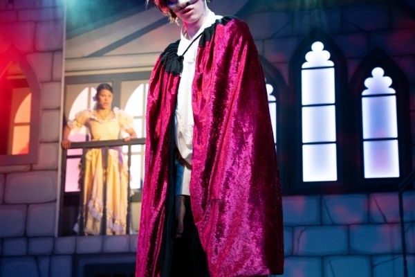 A performer portraying the Beast with a horned mask skulks across the stage, with Belle in a yellow dress visible in the background beside a castle backdrop.