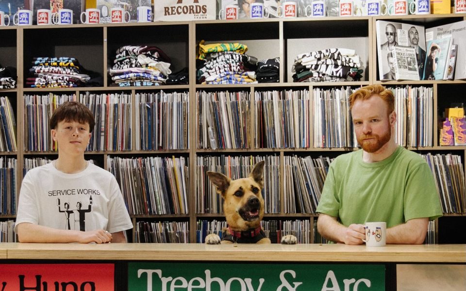Two people and a dog stand behind a counter in front of a large display of vinyl records on shelves.