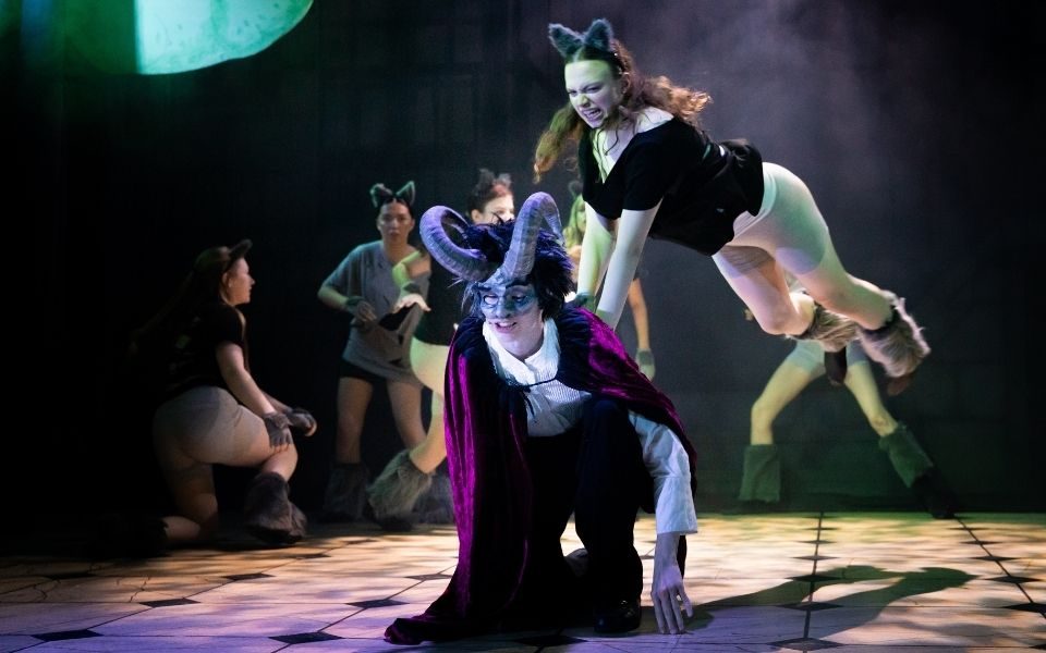 A performer portraying the Beast in a horned mask and cape is being attacked by a group of wolves, played by young people in furry footwear and ears.