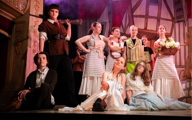 A group of young performers stand together on stage, wearing aprons and dresses. One holds a basket of flowers and another has sausages looped around their neck. Gaston stands proudly beside the group, resting a prop shotgun on their shoulder.
