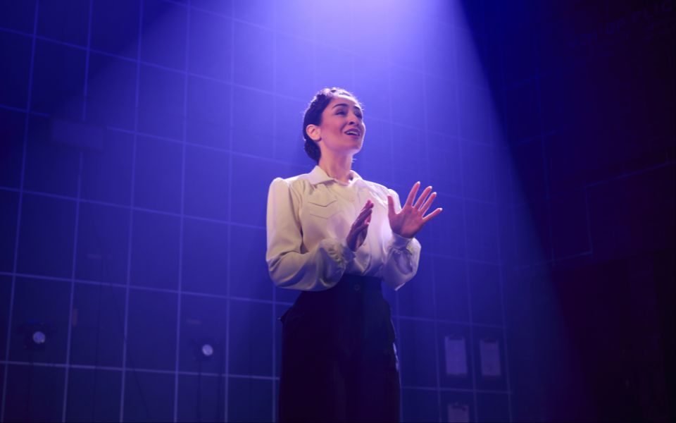 A performer in a blouse and skirt sings beneath a blue-purple spotlight on stage.