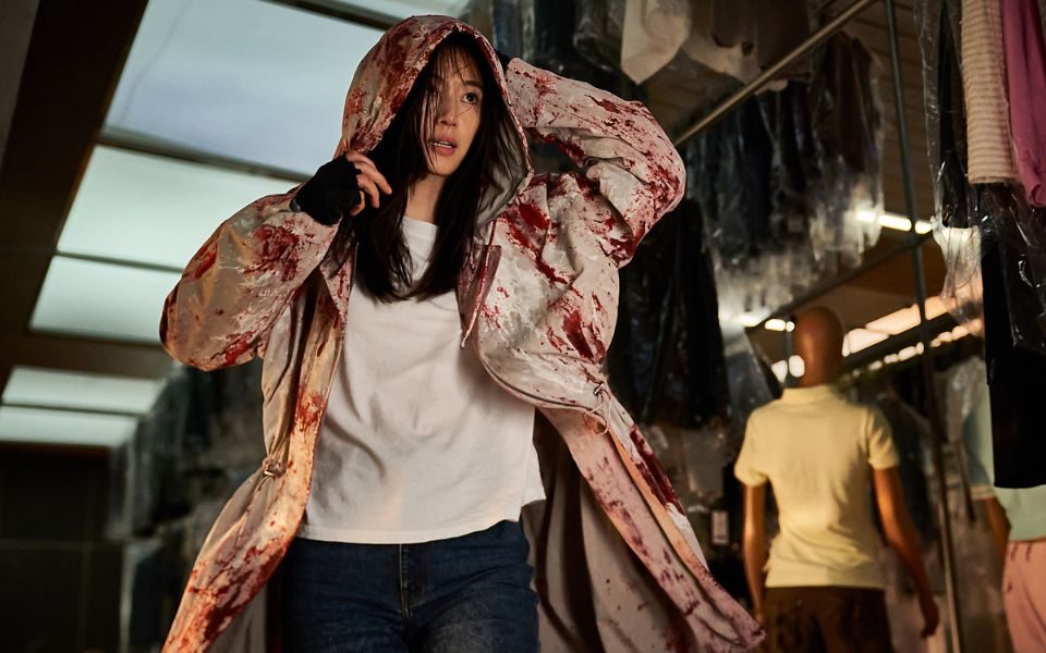 A young woman walks through a corridor featuring clothing rails and mannequins. She is wearing a blood-stained overcoat.