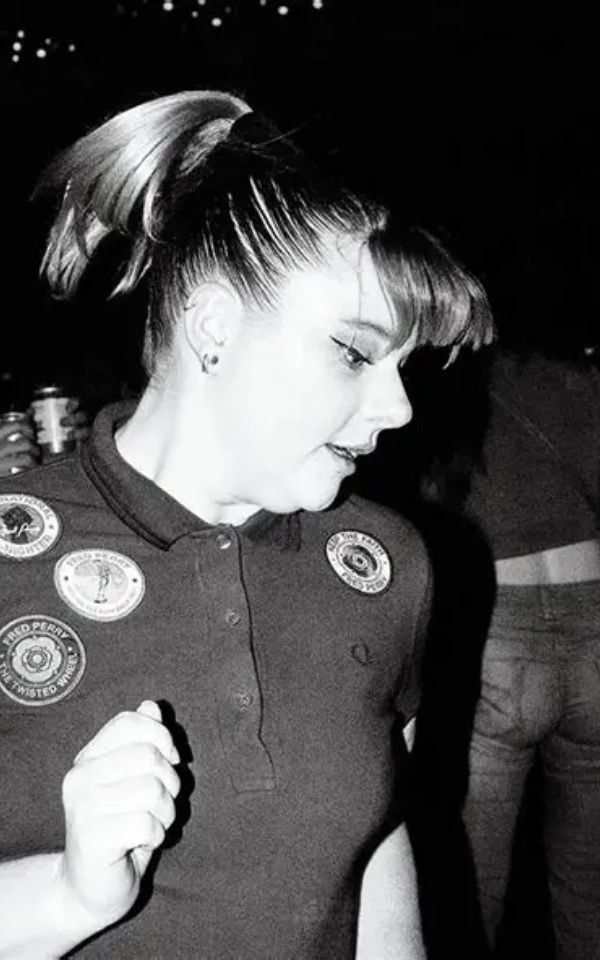 A girl with a ponytail and polo shirt with patches dances in a club.