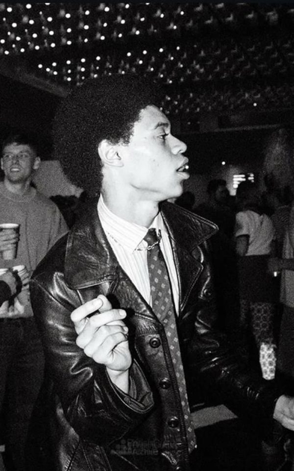 A young man in a shirt, tie and leather jacket dances in a crowded dancefloor in a club.