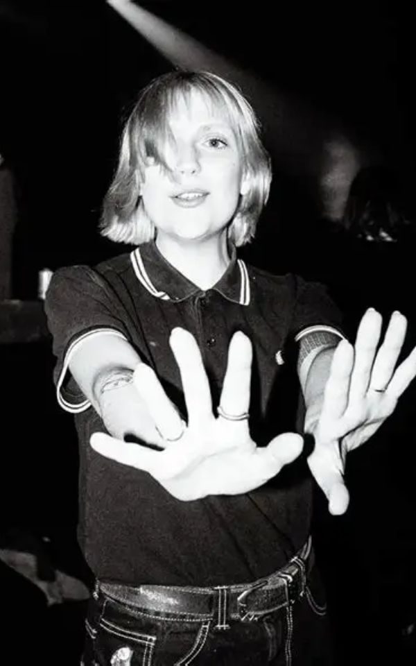 A girl with shoulder length hair in a Fred Perry polo shirt candidly smiles and puts her hands up to the camera.