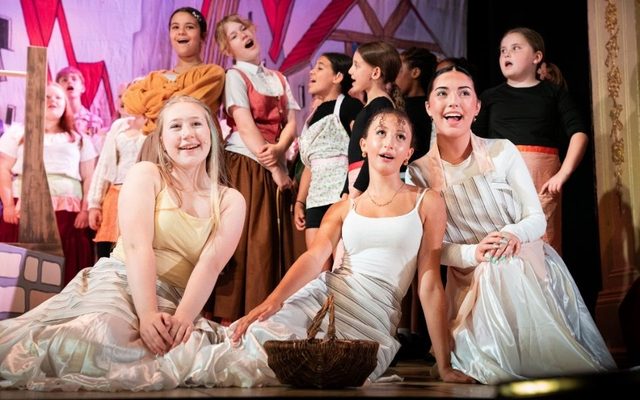 Three young performers smile and sing together, knelt at the front of the stage with a wicker basket in front of them. A group of young people sing behind them in the background.