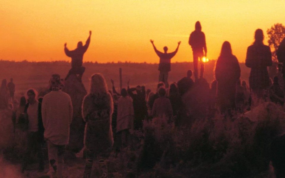 The silhouettes of a group of people on a field facing an orange sunset. Some of them are stood on rocks and raise their arms in the air.