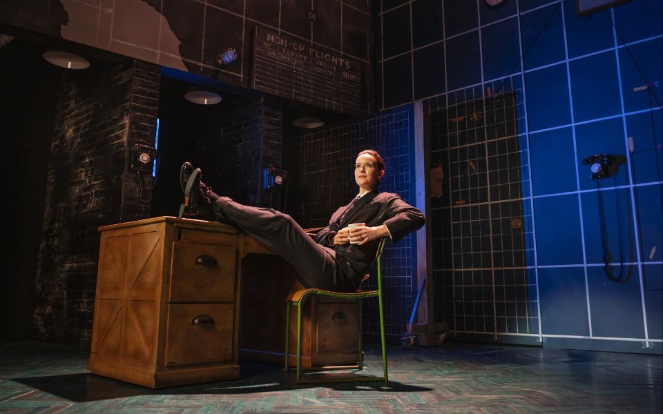 A suited performer sits back in a chair holding a mug, with legs crossed casually on a wooden desk.