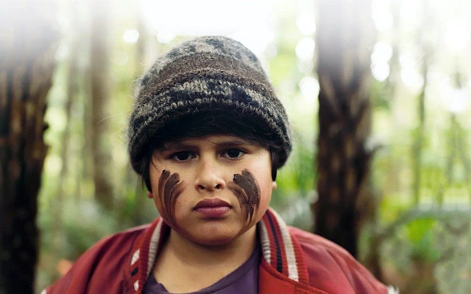 Hazy close-up of a young boy with mud on his face in the style of war paint, stood in a forest looking at the camera.