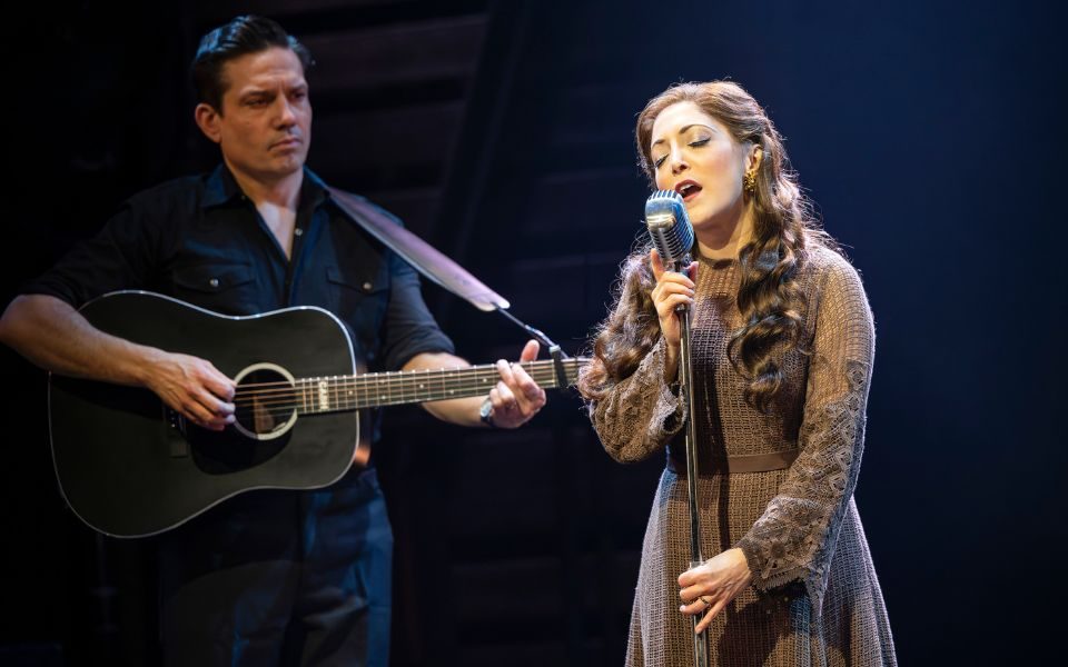 Christina Bianco playing June Carter Cash singing into a microphone while Christopher Ryan Grant playing Johnny Cash plays guitar in the background.