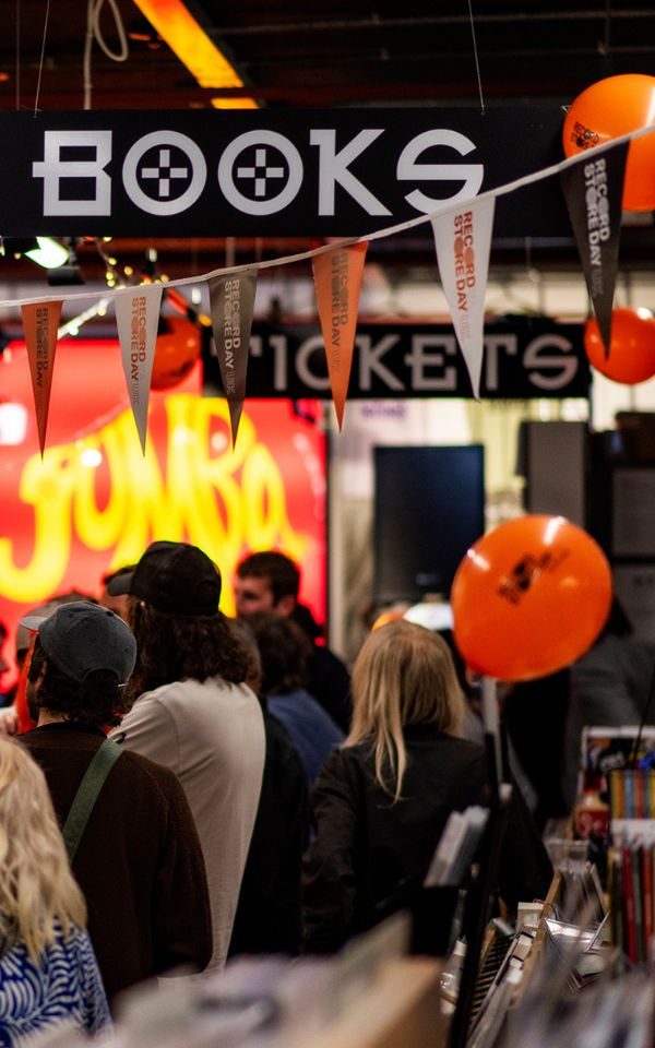 A crowd of people visible from behind, lined up in a record shop and facing a bright sign reading 'Jumbo'. There is bunting in the foreground and hanging signs reading 'Books' and 'Tickets'.