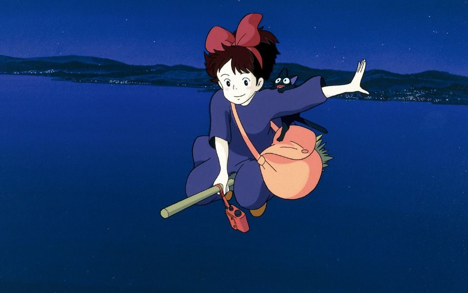 Animated still in anime style. A girl rides a broomstick over a deep blue ocean through a night sky. She has a red bow in her hair, a pastel pink satchel and a black catch clinging to her shoulder.