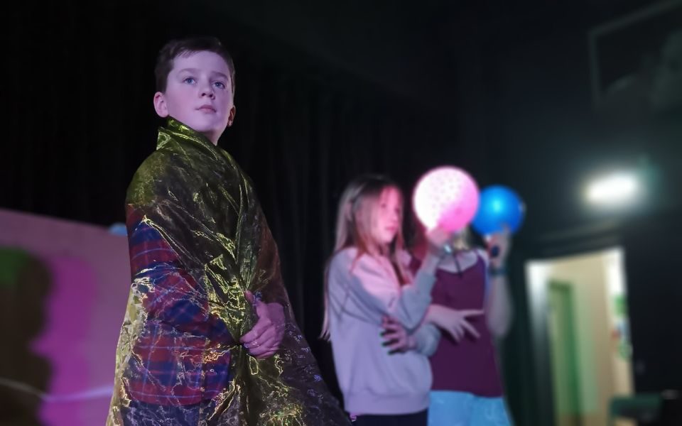 A boy draped in translucent fabric looks off camera, beside two other young people holding light-up pink and blue balloons.