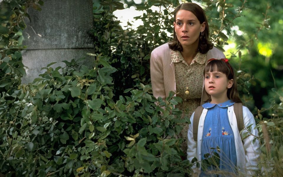 Amongst some bushes, a woman and a child look at something in the distance. The child has a red ribbon in her brown hair and wears a backpack.