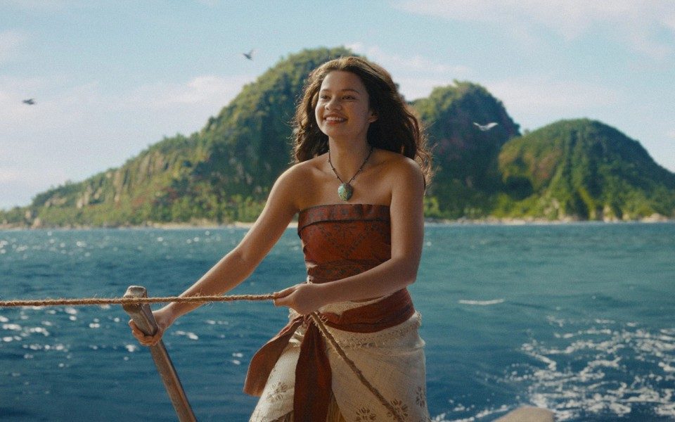 A girl in a patterned red top and skirt smiles as she holds a rope and an oar, moving across a tranquil ocean with greenery in the background.