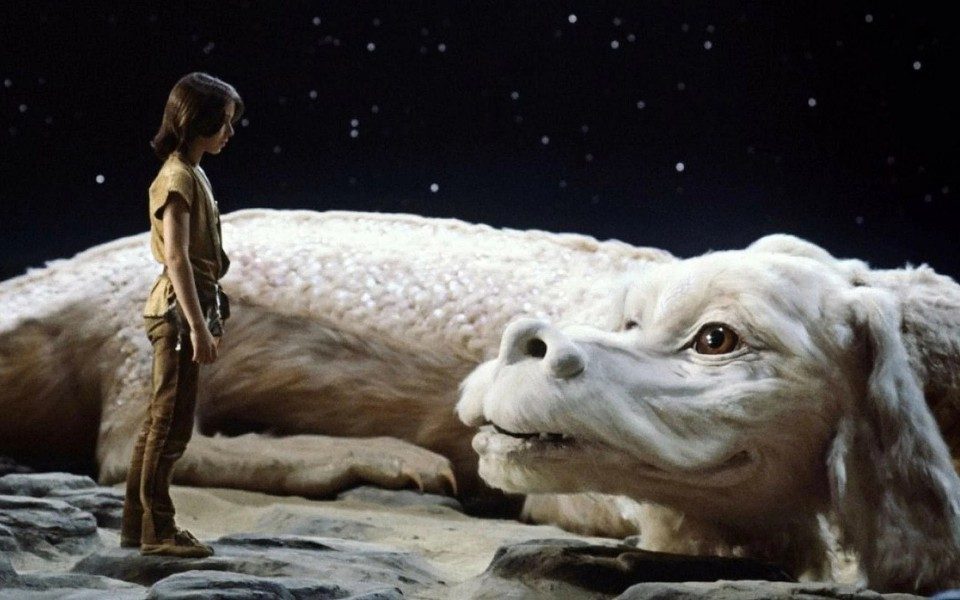 Against a starry night sky, a small child stands on a rocky ground facing a large puppet of a furry, white, dragon-like dog creature with large brown eyes.