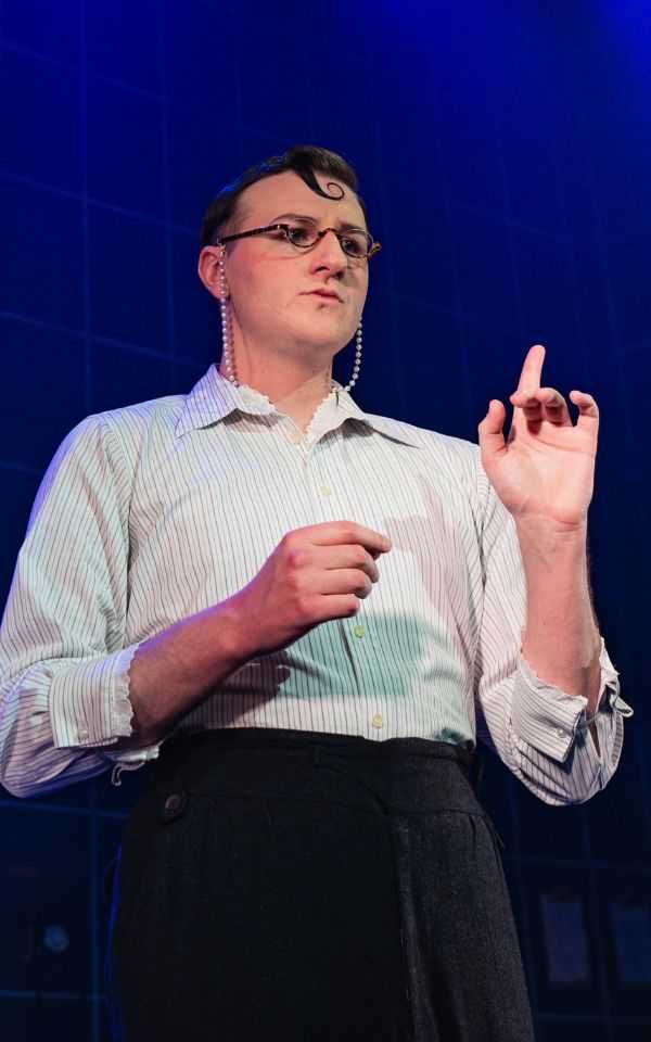 A performer wearing glasses and a white shirt raises a finger.