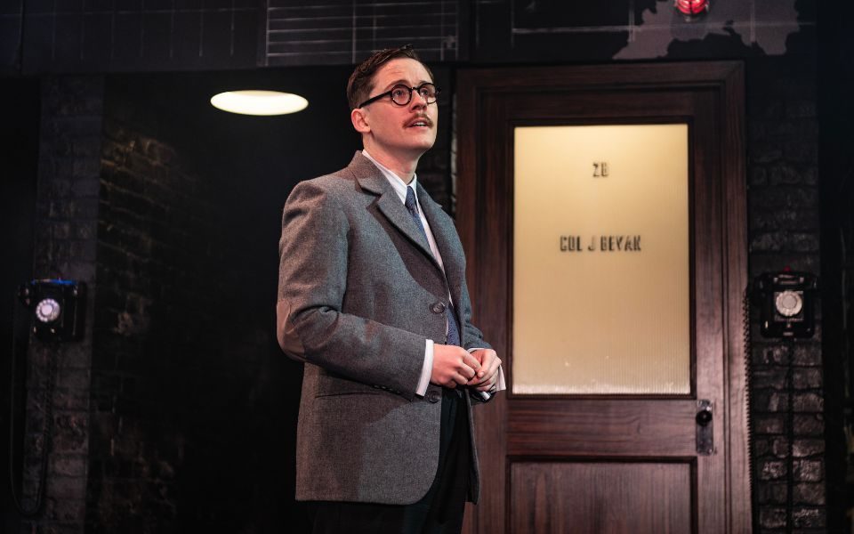 A performer in glasses and a period suit stands on a stage in front of an office door and vintage telephones.