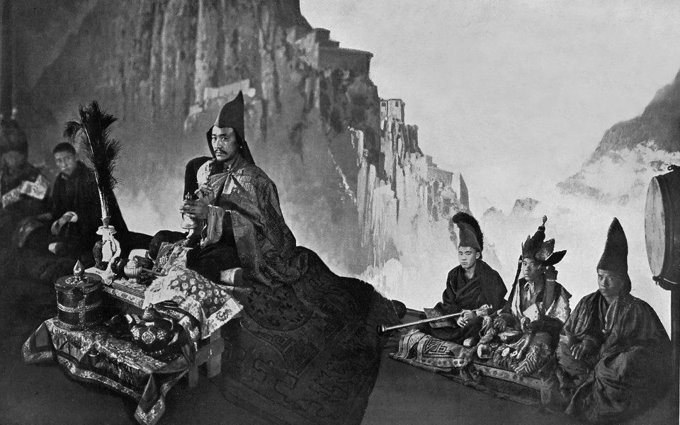 Black and white film still. A man in a pointed hat and long roe sits above an elaborately laid table of trinkets, alongside other men in robes. They sit in front of a grand clifftop landscape.