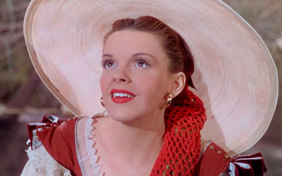 Judy Garland looks upwards with a smile, wearing a white wide-brimmed hat, red dress and red lipstick.