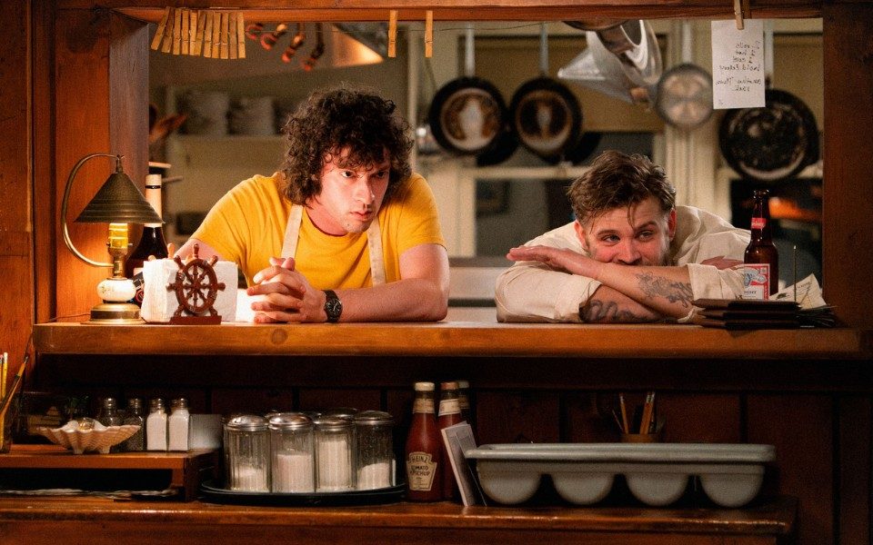 Two chefs lean on a counter as they look out from a kitchen laden with pots and pans. Restaurant cutlery and condiments are stacked in the foreground.