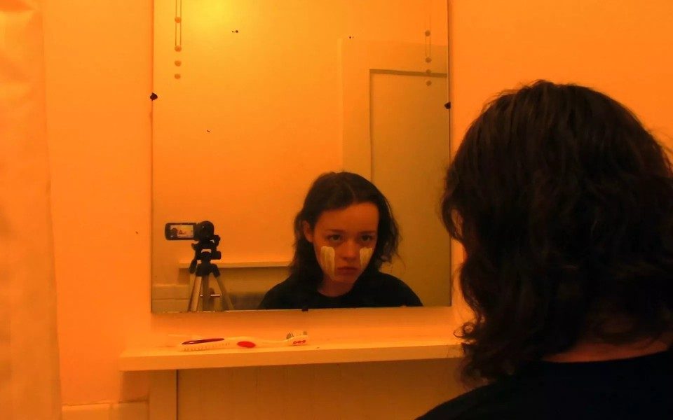 In a yellow-tinged bathroom, a girl with white face paint across her cheeks looks into the mirror with a vacant gaze, beside a digital camera on a tripod.