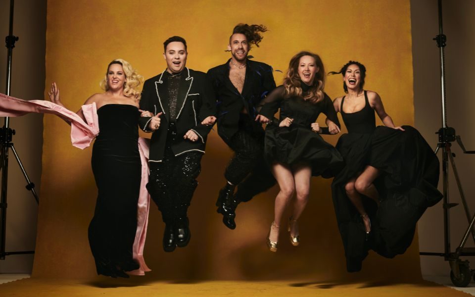 A smiling group of actors wearing black suits and dresses link arms and jump in unison in front of a golden backdrop in a photography studio.
