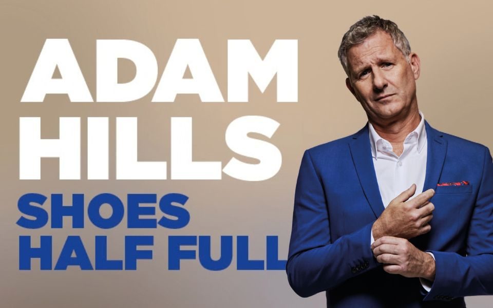 Adam Hills stands wearing a blue suit. Text reads Adam Hills. Shoes Half Full.