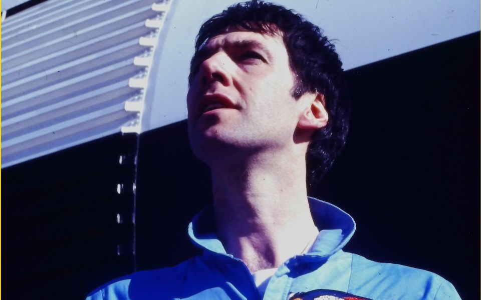 Clint Boon in blue overalls and a patch with a logo of a spaceship. He looks up, off-camera, standing in front of a white and black rocket.