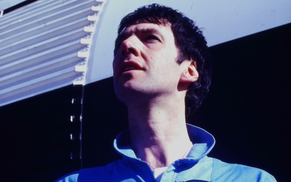 Clint Boon in blue overalls and a patch with a logo of a spaceship. He looks up, off-camera, standing in front of a white and black rocket.