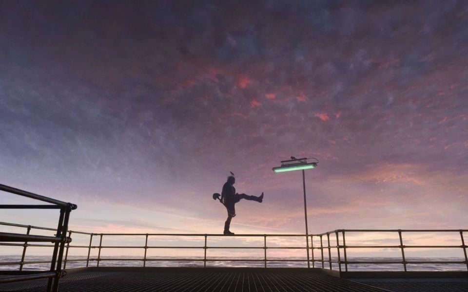 The silhouette of a man high-kicking and holding a spade, on top of some metal railings in front of a scenic coastal backdrop and purple sky.