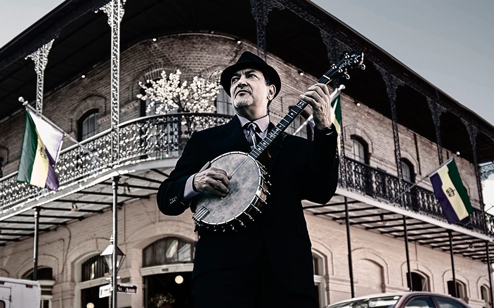 Shot from below, Don Vappie stands at a street corner, playing a banjo and looking into the distance, wearing a black suit and pork pie hat.