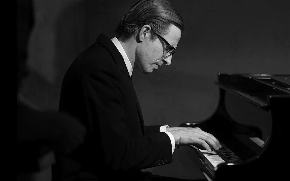 Black and white still of a man in a black suit, glasses and combed hair playing the piano.