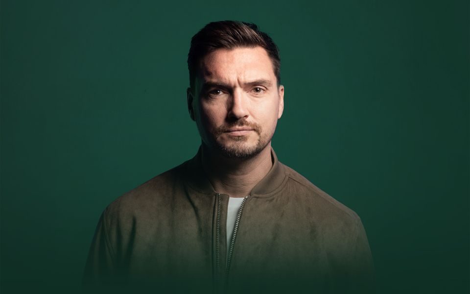 Against a dark green backdrop, Jack Skipper stands in a brown jacket and holds a neutral expression to the camera.