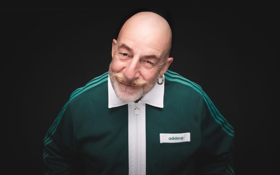 Jeff Innocent, a bald man with a beard and moustache, leans towards the camera, smirking. He wears a dark green Adidas tracksuit with a white collar.