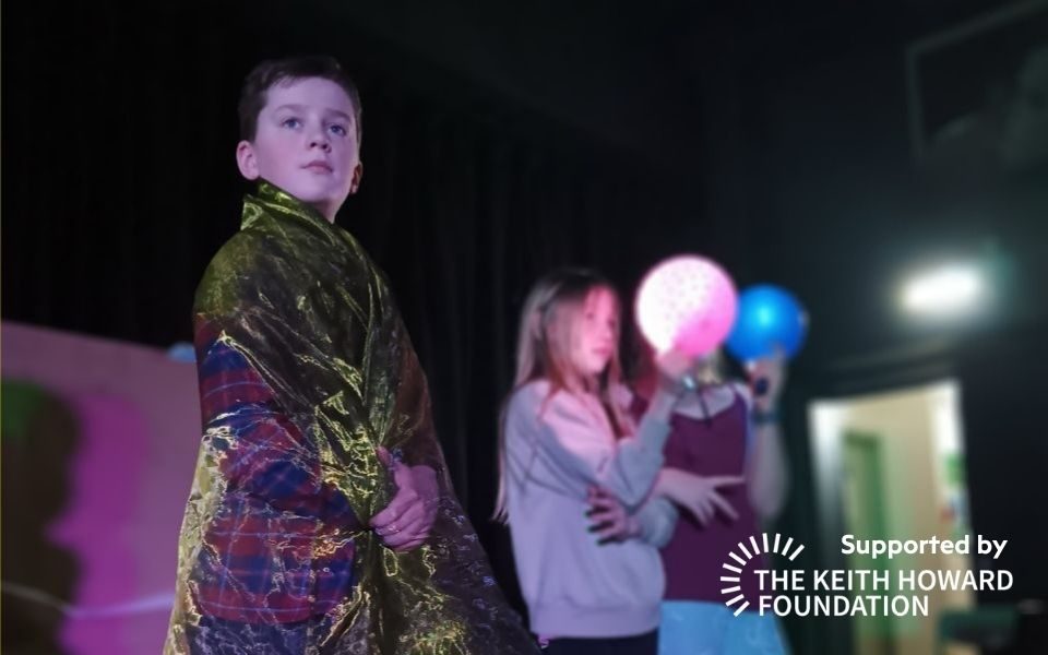 A boy draped in translucent fabric looks off camera, beside two other young people holding light-up pink and blue balloons. Text reads: Supported by the Keith Howard Foundation.