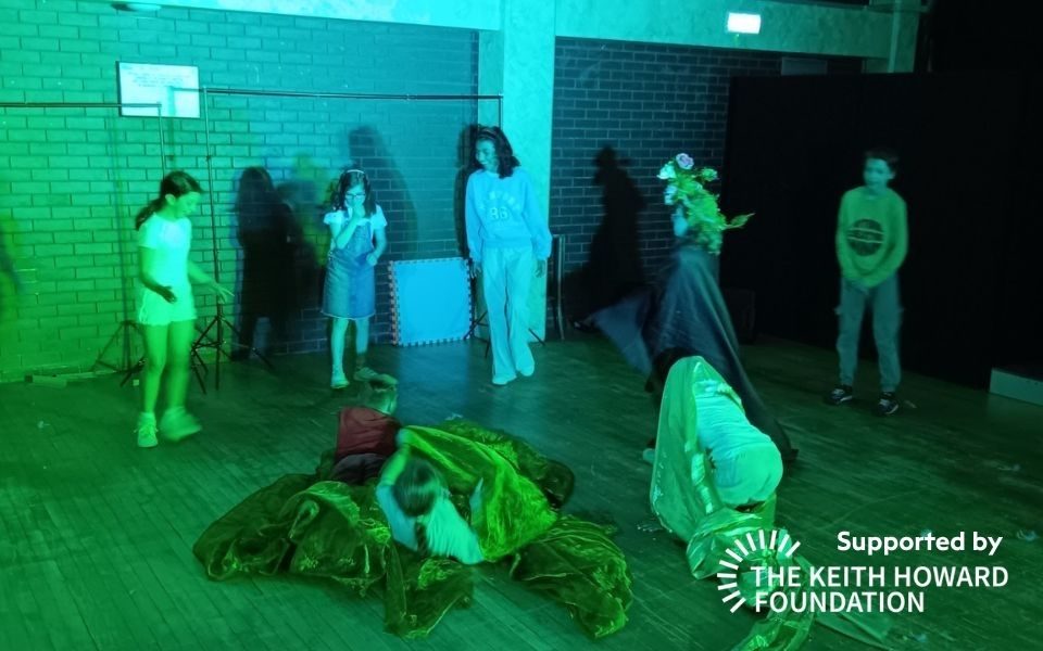 A group of children in a studio setting playing with sheets of fabric beneath a green light. Text reads: Supported by the Keith Howard Foundation.