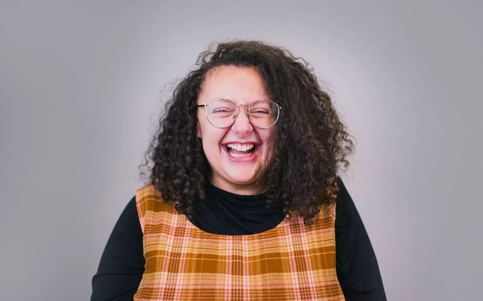 Close up of a woman in glasses with shoulder-length curly hair laughing in front of a blank backdrop.