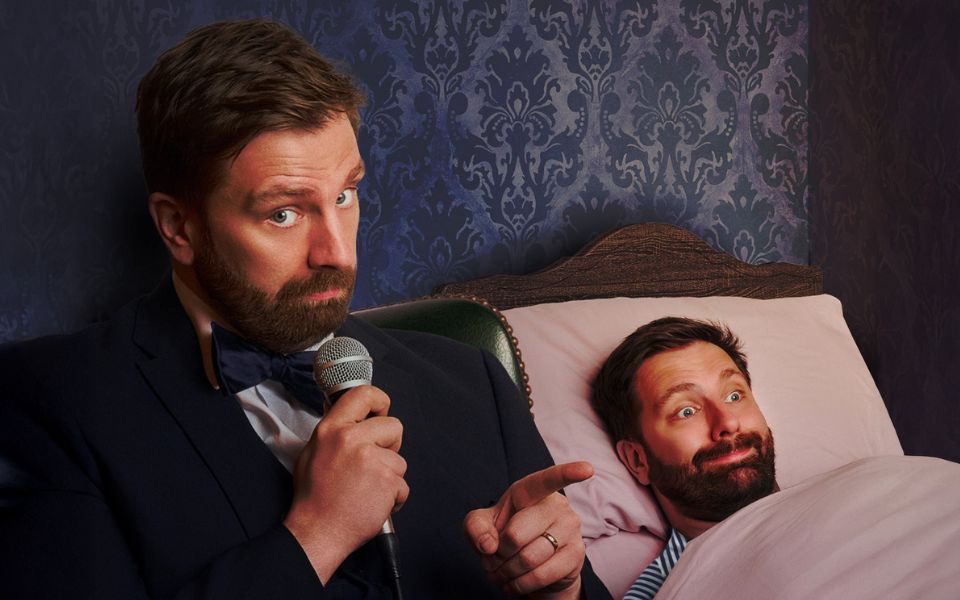 Pierre Novellie sits beside a bed, holding a microphone. In the bed, a second Pierre Novellie is tucked in beneath a pink blanket with a comical smile.