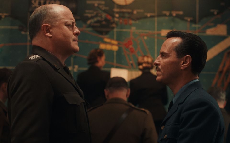 Two men stand face-to-face with stern expressions, both wearing different military uniforms. They are in a war room, and a map and other people in period costumes are visible in the background.