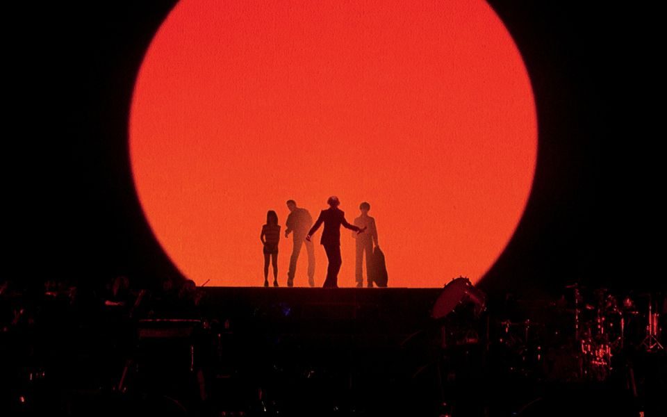 Silhouettes of four band members posed on stage, in front of a large blood orange circular light,