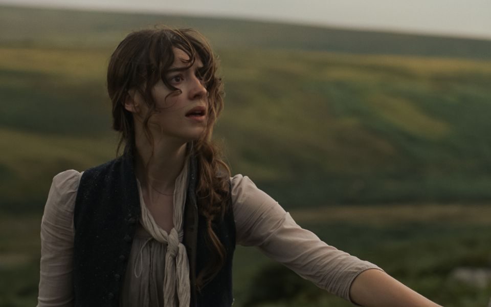 In a misty countryside setting, a woman with curly windswept hair and period clothing looks off-camera.