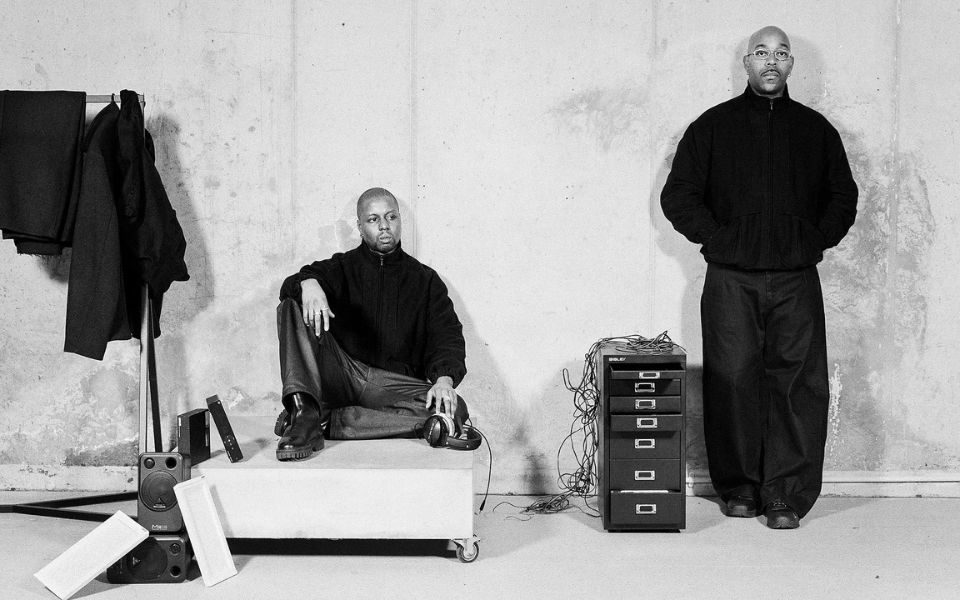 A man sits on a crate in a studio warehouse setting, next to speakers, headphones and a metallic cabinet. Another man stands with his hands in his jacket pockets, wearing baggy trousers.